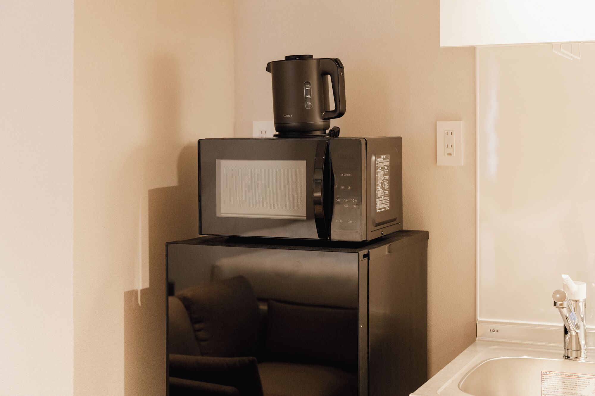 Una cocina compacta con microondas y cafetera sobre un mueble, un lavabo y una silla.