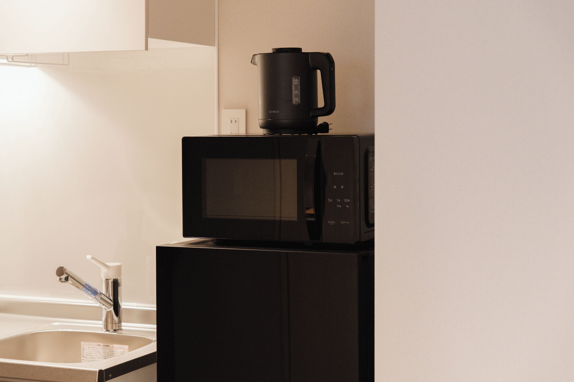 A kitchen with a microwave and a coffee maker on top, a sink with a faucet, and a wall-mounted light.