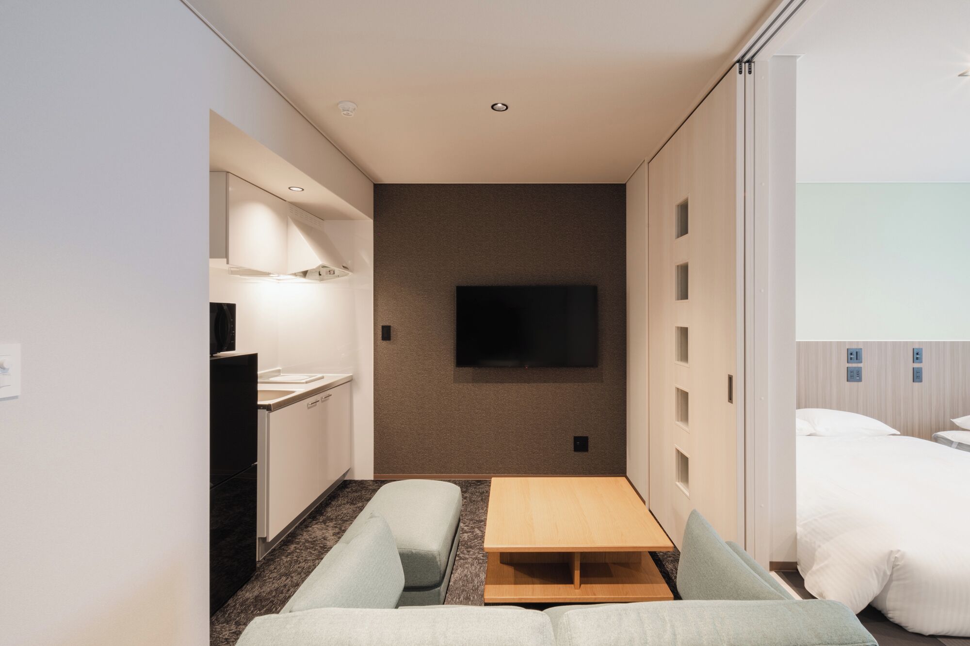 Una habitación de hotel compacta con kitchenette, un televisor de pantalla plana montado en la pared, una cama y una pequeña mesa de madera.