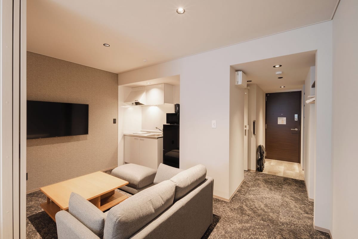 Ein modernes Hotelzimmer mit einer Couch, einem kleinen Tisch, einem an der Wand montierten Fernseher und einer Küchenzeile.