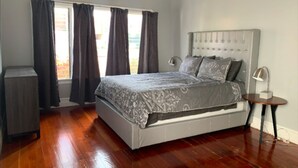 A bedroom with a large bed, a bedside table, a lamp, and a window with curtains. - High Ceiling, Cozy 1BR Apt, Walk to Metro & Shops (Pasadena)