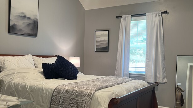 Ein Schlafzimmer mit einem Bett, einem Deckenventilator, einem Fenster mit Vorhängen und einem an der Wand befestigten Spiegel.