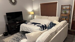 Ein Schlafzimmer mit einem Bett, einem Fernseher, einer Lampe, einem Fenster mit Jalousien und einem Regal mit einer Pflanze.