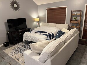 A bedroom with a bed, a television, a lamp, a window with blinds, and a shelf with a plant. - Charming House near Ole Miss  (Oxford)