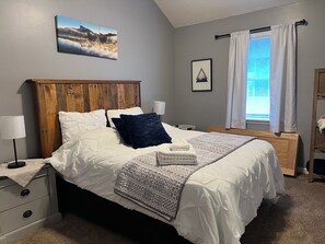 A bedroom with a wooden headboard, a bed with white bedding and a patterned throw, a bedside table with a lamp, and a window with curtains.