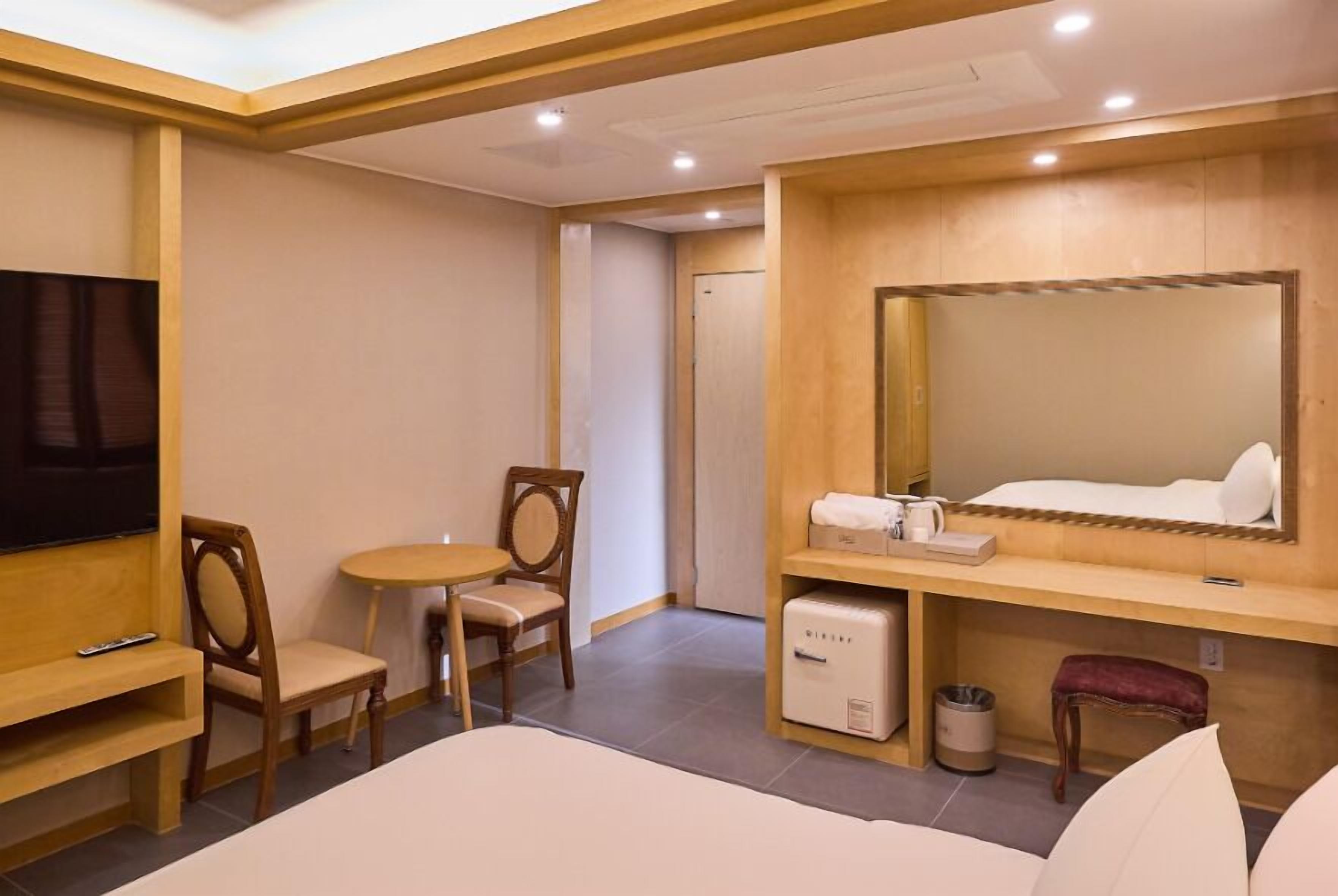Habitación de hotel con cama, televisión, escritorio con silla, mesita pequeña, espejo y refrigerador minibar.