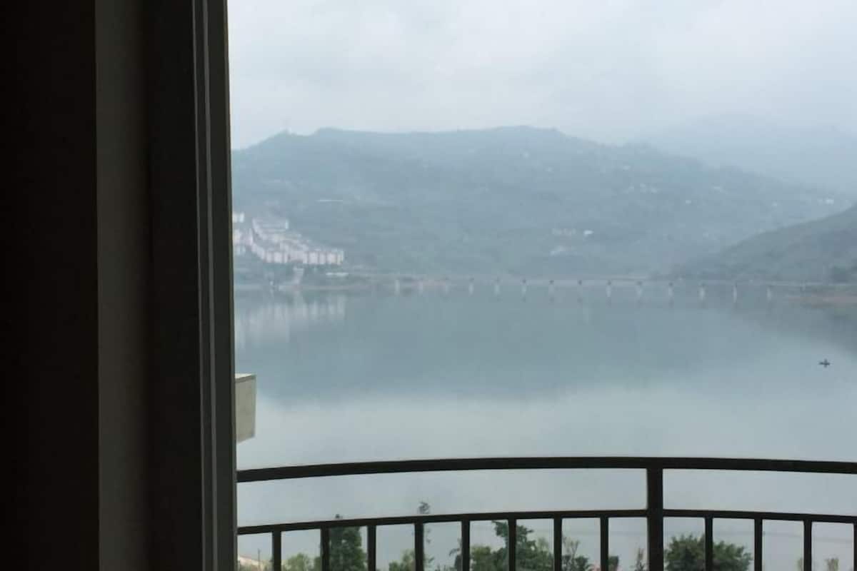 Vista desde un balcón sobre un lago tranquilo, con montañas a lo lejos y un puente en el centro.