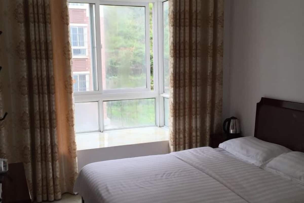 A hotel room with a bed, curtains, a window, and a nightstand with a kettle.