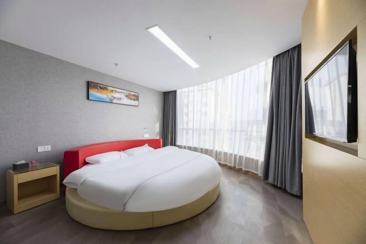 A modern hotel room with a large bed, a nightstand, a painting on the wall, and a large window with curtains.