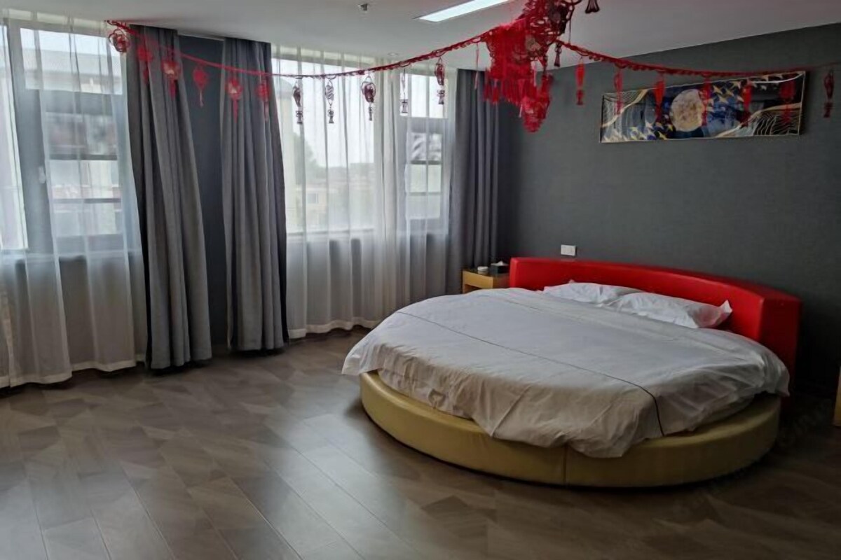 A bedroom with a round bed, grey curtains, a red headboard, and a painting on the wall.