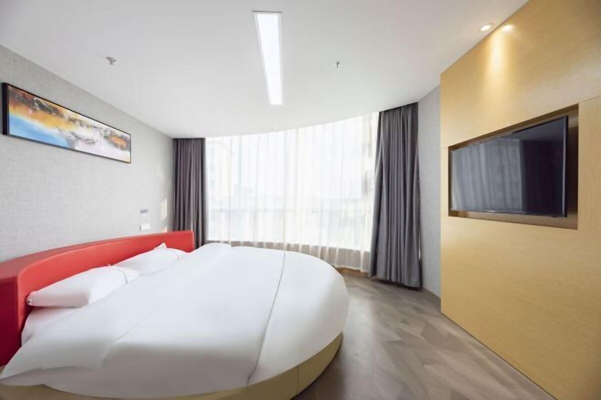 A modern hotel room with a large bed, a flat-screen TV, and a large window with curtains.
