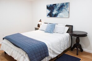 Ein ordentlich bezogenes Bett mit einer blauen Steudecke, einer Nachttischlampe und einem kleinen runden Tisch daneben.