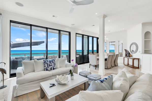 A spacious living room with a large window overlooking a beach, a white sofa, a coffee table, and a dining area.
