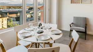 Comedor con una mesa redonda dispuesta para cuatro personas, sillas blancas y vistas a un puerto deportivo y edificios a través de grandes ventanales.