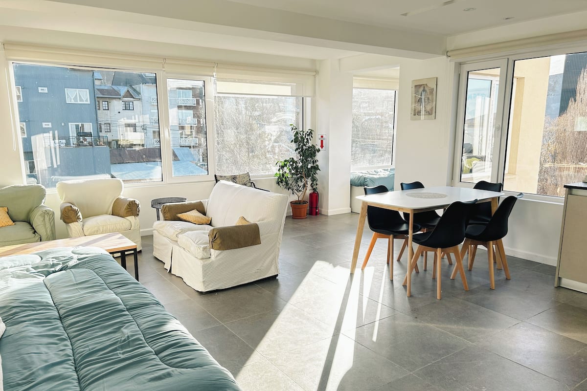 Un salón amplio con sofá, una mesa de comedor con sillas y ventanas grandes que ofrecen vistas a edificios y árboles.