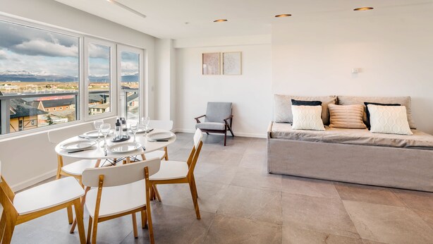 Una sala de estar moderna con comedor, un sofá gris con cojines estampados y vistas a un paisaje montañoso a través de amplios ventanales.