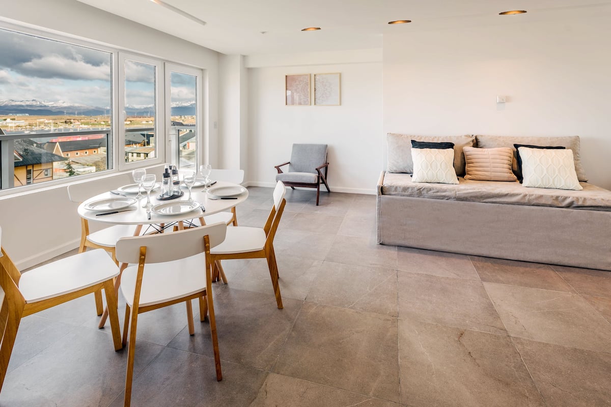 Una sala de estar moderna con comedor, un sofá gris con cojines estampados y vistas a un paisaje montañoso a través de amplios ventanales.