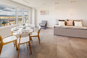Un salon moderne avec un coin repas, un canapé gris agrémenté de coussins à motifs et une vue sur un paysage montagneux grâce à de grandes fenêtres.