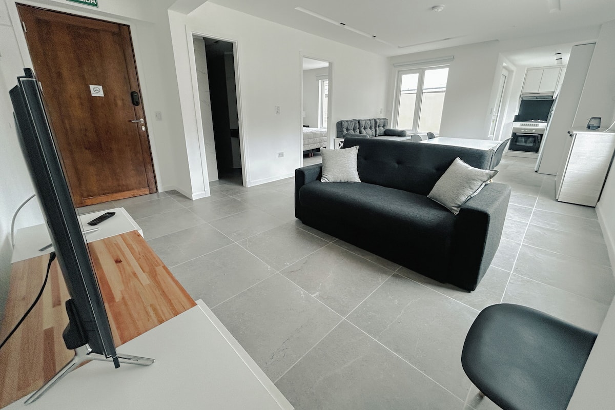 Una sala de estar moderna con un sofá negro, una mesa de centro de madera y un televisor. Se observa un área de cocina al fondo con un refrigerador y una estufa.