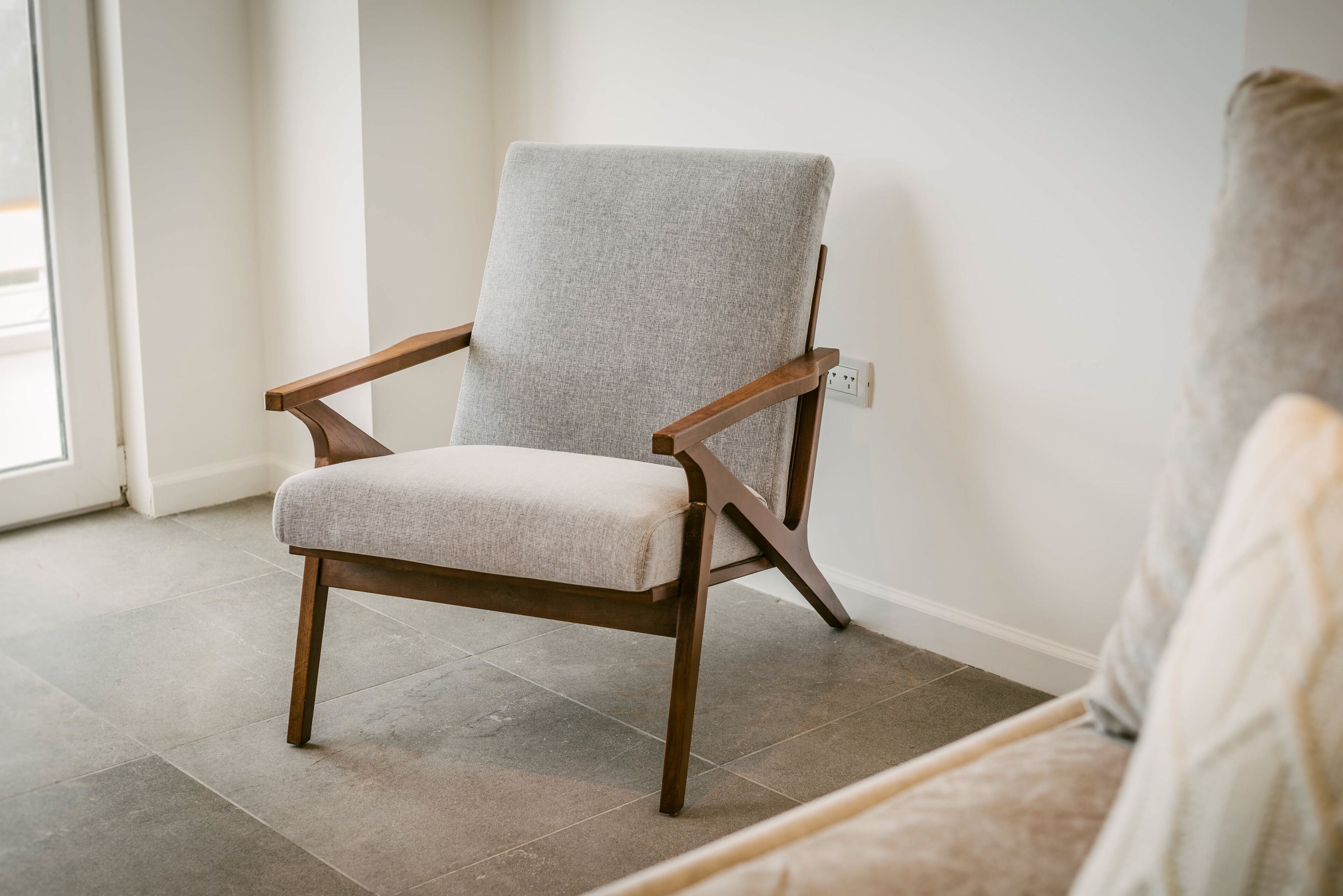 A chair with a light grey upholstered seat and wooden armrests, positioned in a room with a tiled floor and a white wall.
