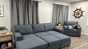 Ein modernes Wohnzimmer mit einem grauen Ecksofa, einem Holztisch, einem kleinen Beistelltisch mit Pflanze und einem an der Wand befestigten Schiffsschwungrad.