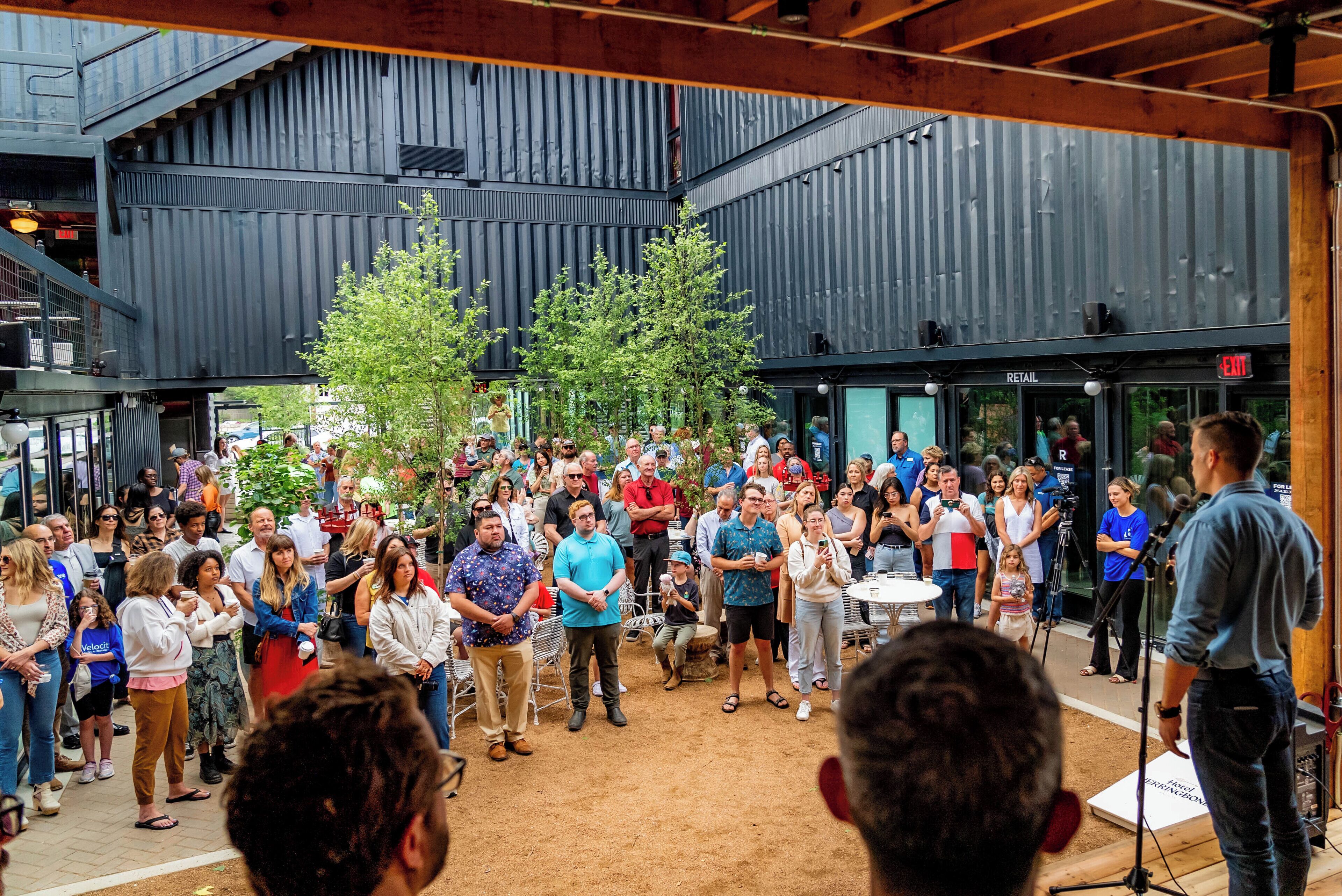 A spacious indoor venue with high ceilings and a modern industrial design, featuring a central seating area surrounded by a crowd.