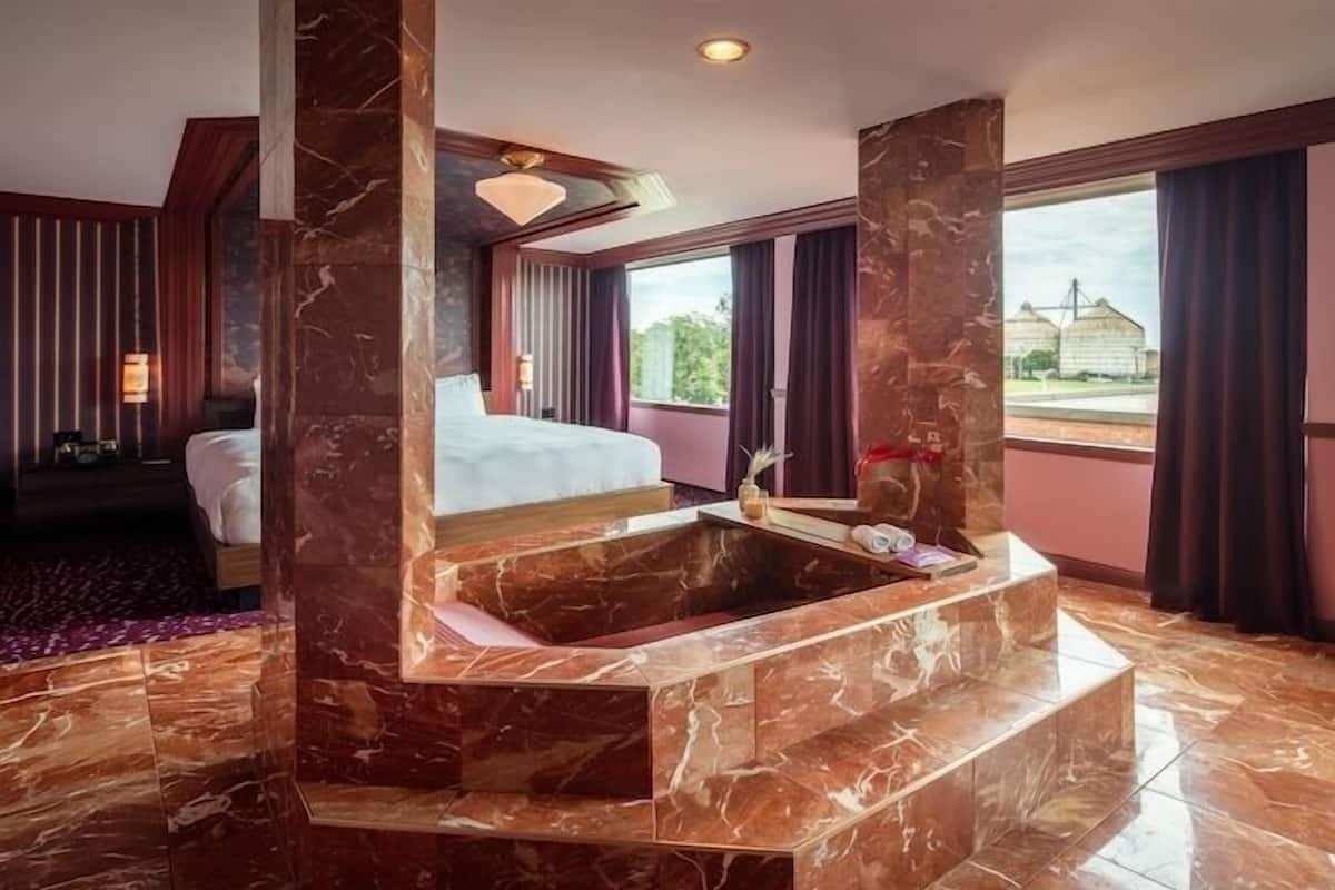 A hotel room with a large bed, a marble bathtub, and a view of the outside.