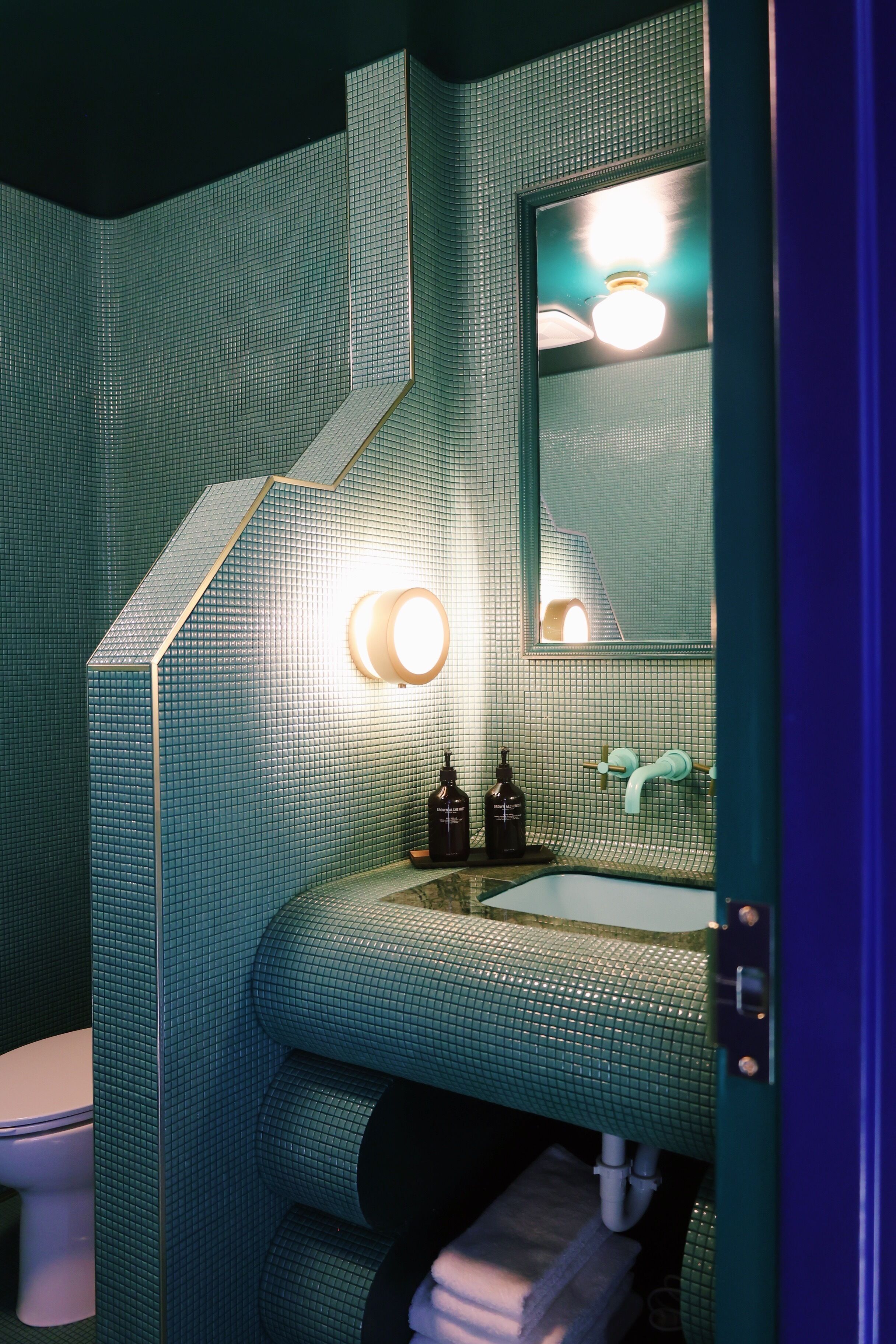 Une salle de bain avec un mur recouvert de carreaux verts, des toilettes blanches, un lavabo avec un miroir et un porte-serviettes.