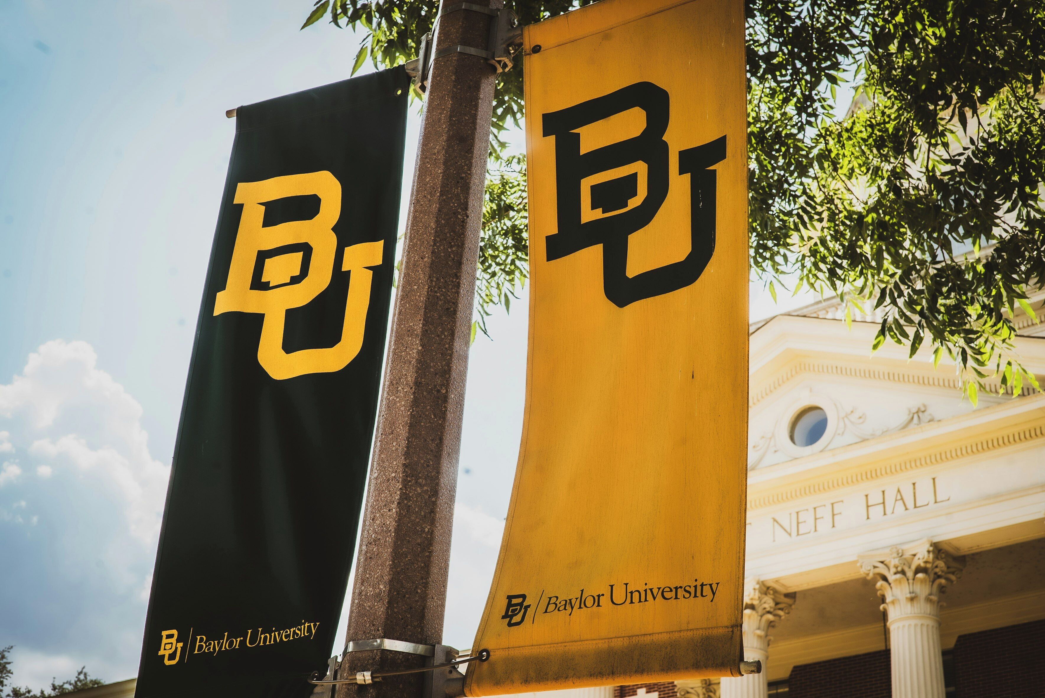 Two Baylor University banners with the university's logo, attached to a pole, with a building labeled "NEFF HALL" in the background.