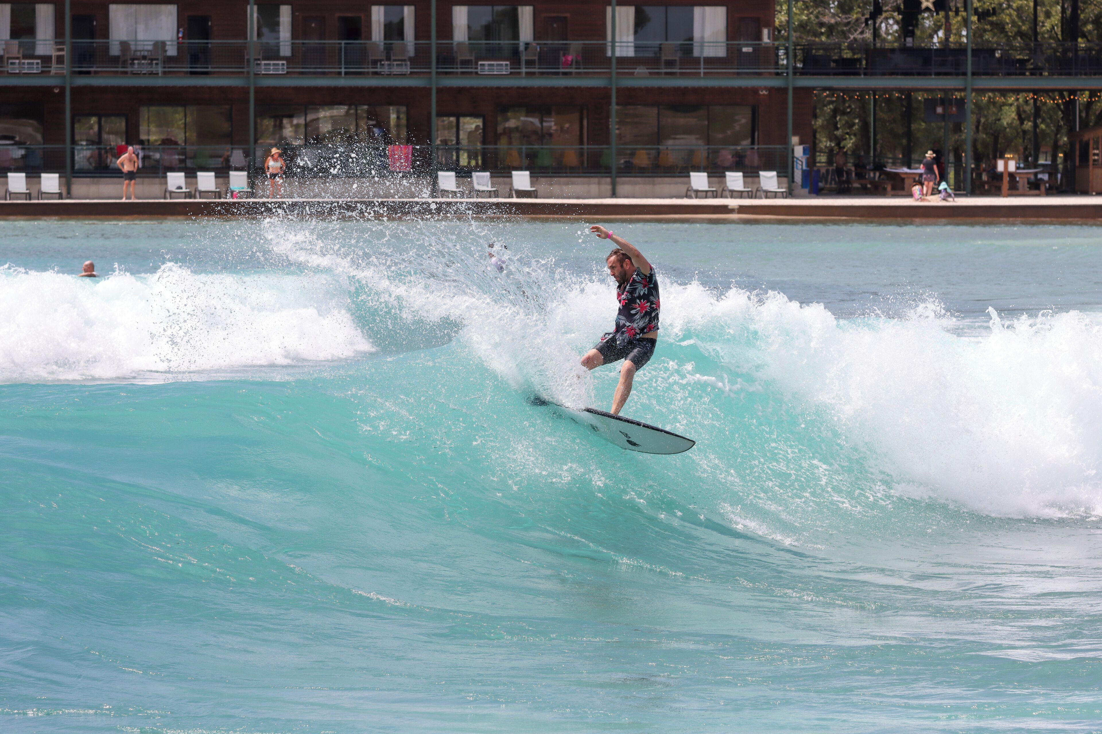 A man surfing on a wave in a pool with a building and lounge chairs in the background.