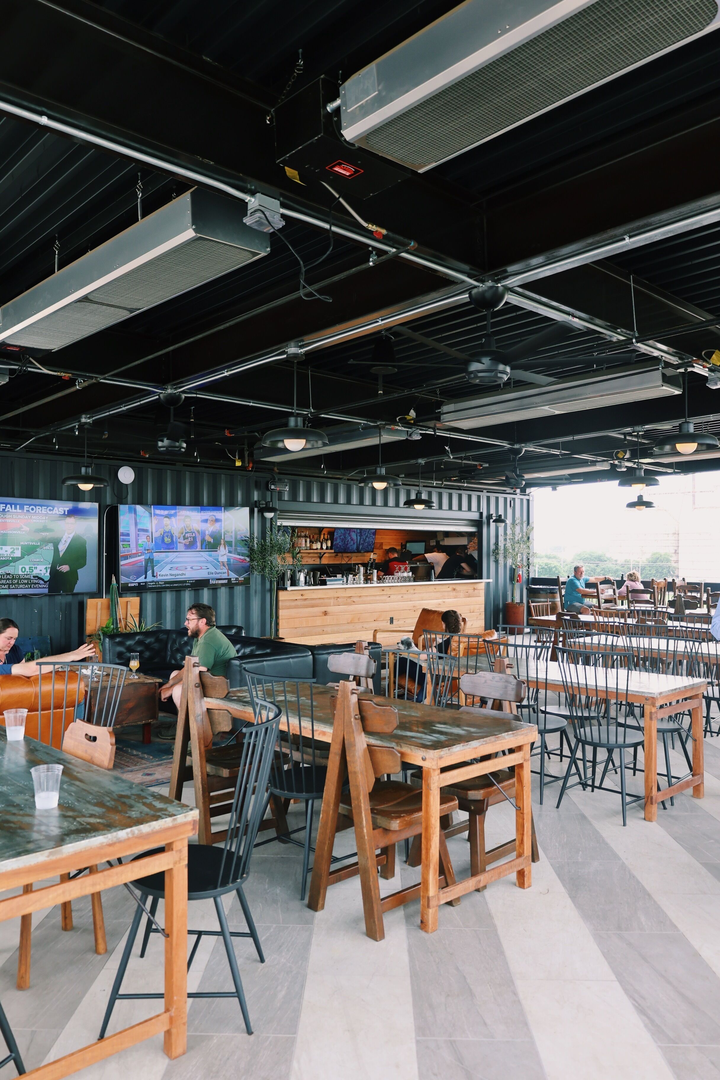 A modern restaurant with wooden tables and chairs, a bar area, and large screens displaying content.