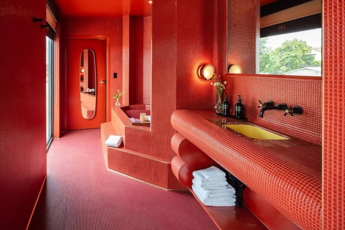 A modern bathroom with a red tiled wall, a large mirror, a yellow sink, and a staircase leading to a shower area.