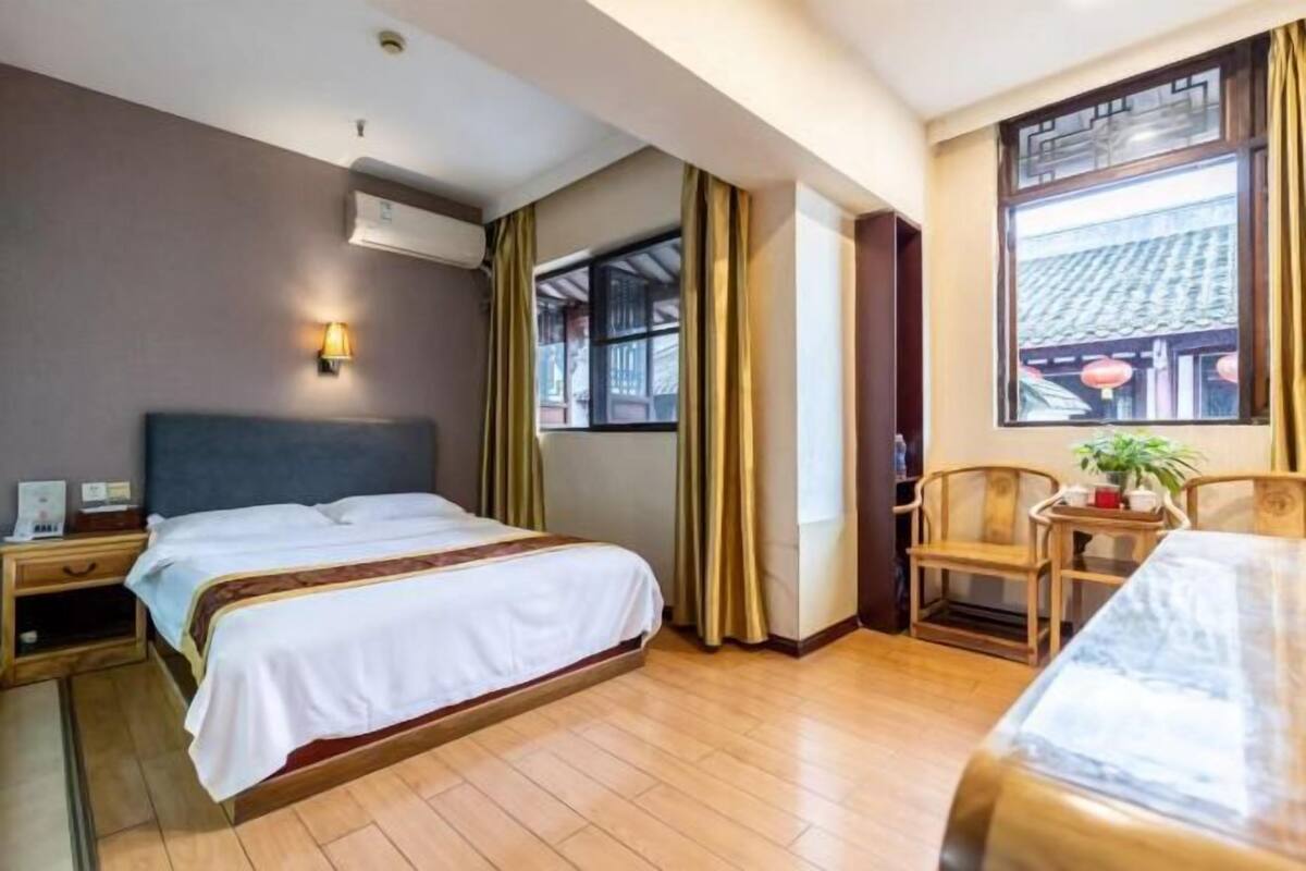 Ein Hotelzimmer mit Bett, Nachttisch, Lampe, Fenster mit Vorhängen und Blick auf traditionelle Architektur.