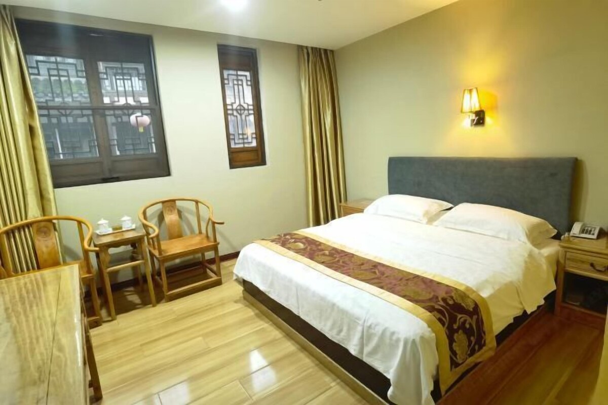 A hotel room with a bed, a bedside table, a lamp, a chair, a small table, and a window with curtains.