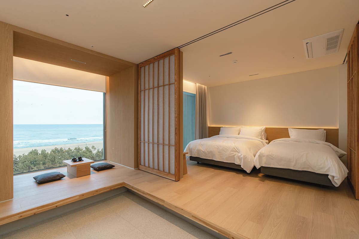 Un dormitorio moderno con un ventanal que ofrece vistas a la playa, una cama con ropa de cama blanca y un suelo de madera.