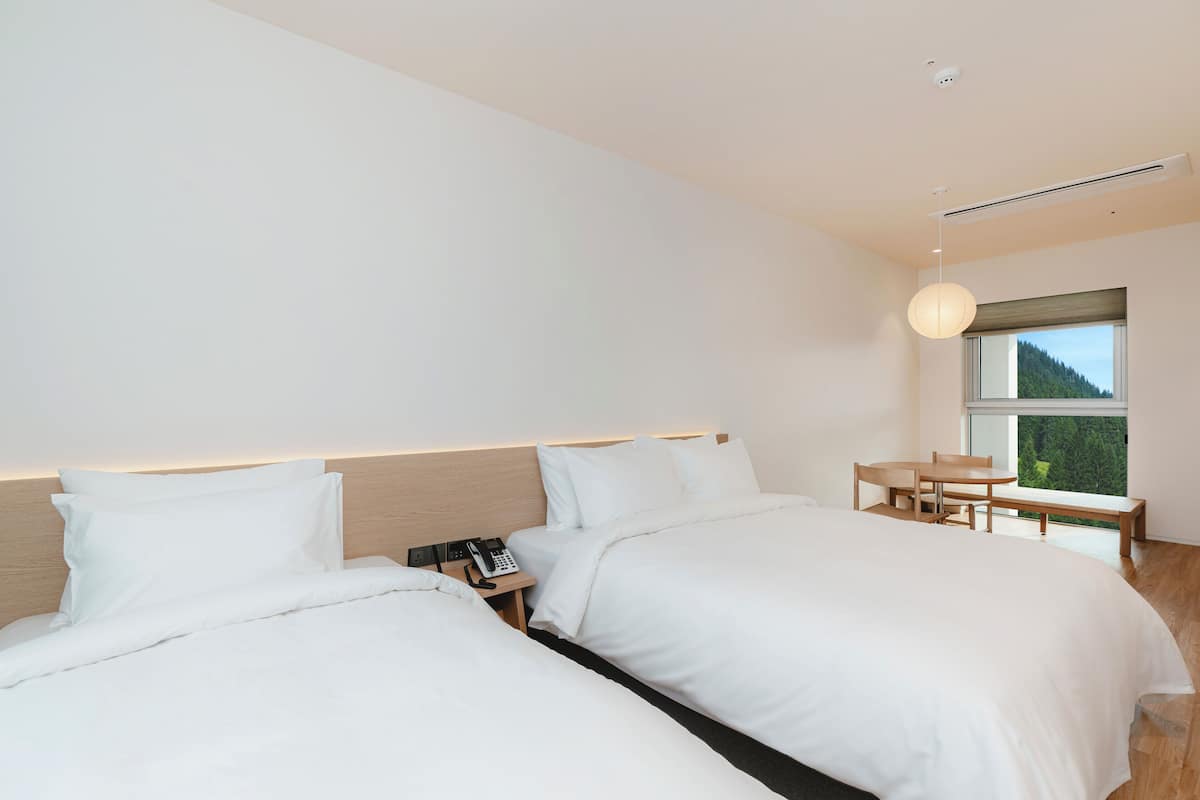 Una habitación de hotel moderna con dos camas, una mesita de noche con teléfono, un escritorio pequeño con silla y una ventana con vistas a zonas verdes.