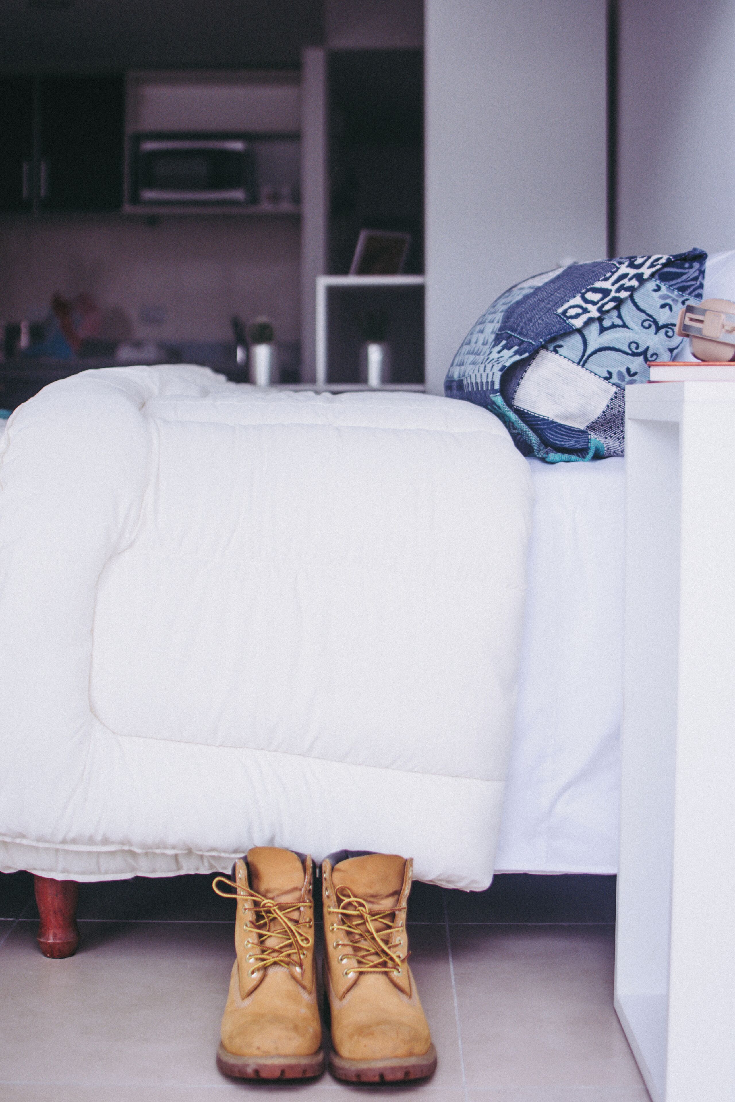 A bed with a white comforter and a pair of brown boots on the floor.