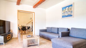 Ein Wohnzimmer mit einer Couch, einem Couchtisch und einem Fernseher. An der Wand hängt ein Gemälde und im Hintergrund befindet sich ein Essbereich.