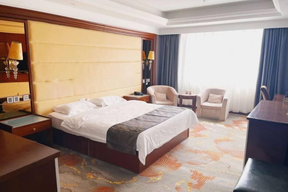 A hotel room with a large bed, two bedside tables, a desk, a chair, and a large window with curtains.