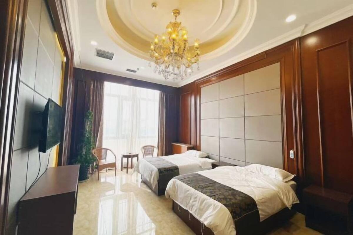 A hotel room with two beds, a TV, a chandelier, and a large window.