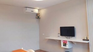 Quarto com televisão de tela plana presa na parede, prateleira branca embaixo e um aparelho de ar condicionado na parede.