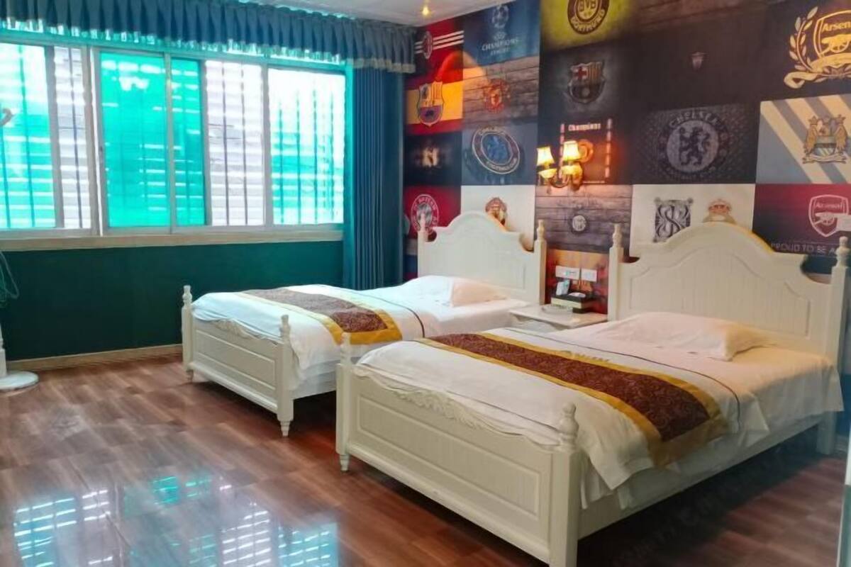 A room with two beds, a large window with blinds, and a wall decorated with sports team logos.