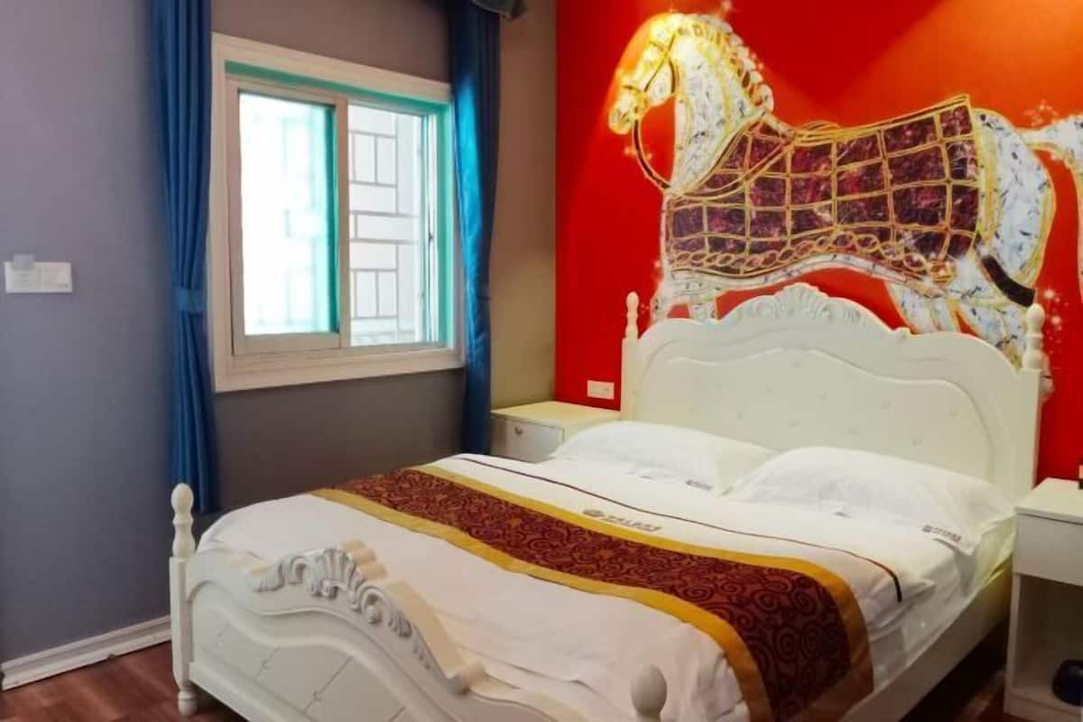 A bedroom with a white bed, a red wall, and a window with blue curtains.