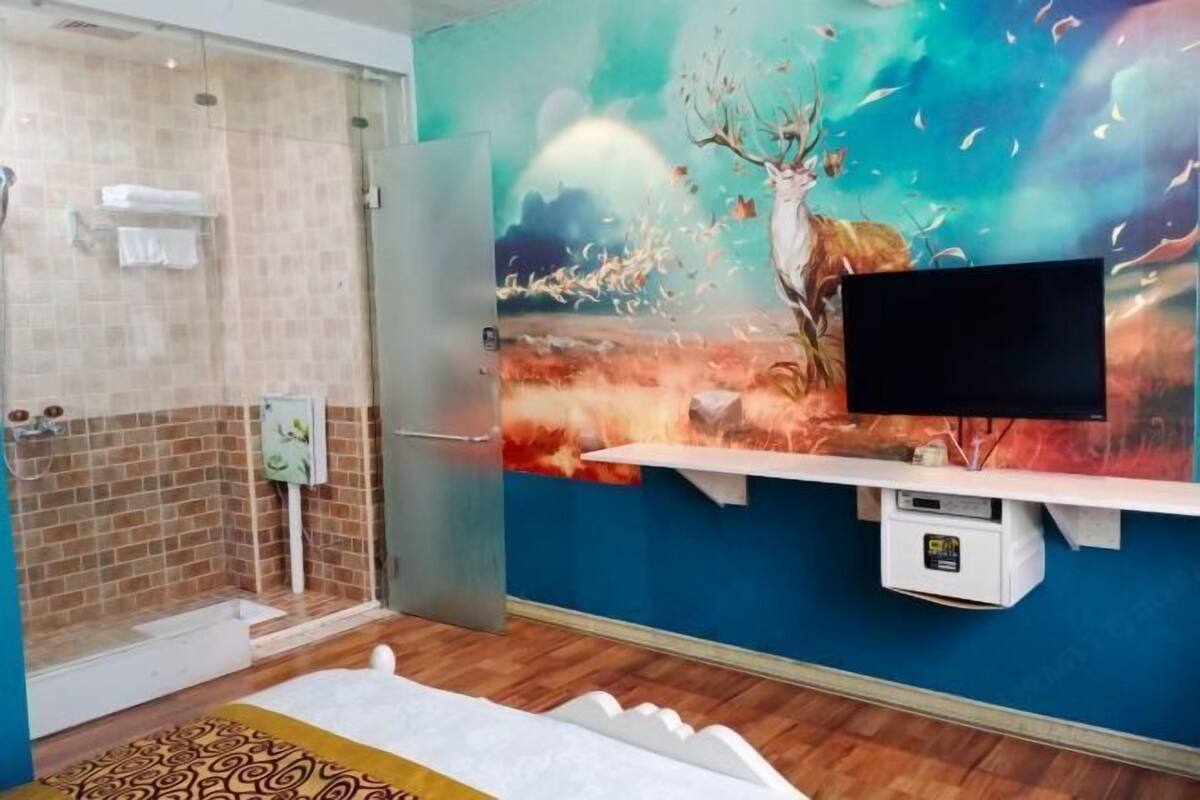 A room with a shower, a bed, a TV mounted on the wall, and a mural of a deer.