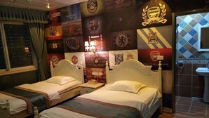 Quarto de hotel com duas camas, uma janela grande com cortinas, parede decorada com logotipos de clube de futebol e banheiro com pia e espelho.