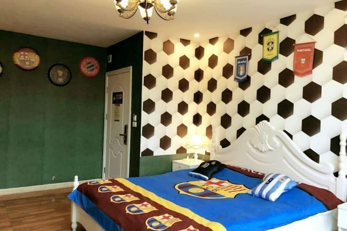 A hotel room with a bed featuring a blue and red blanket with a sports team logo, a white headboard, and a wall with a geometric pattern.