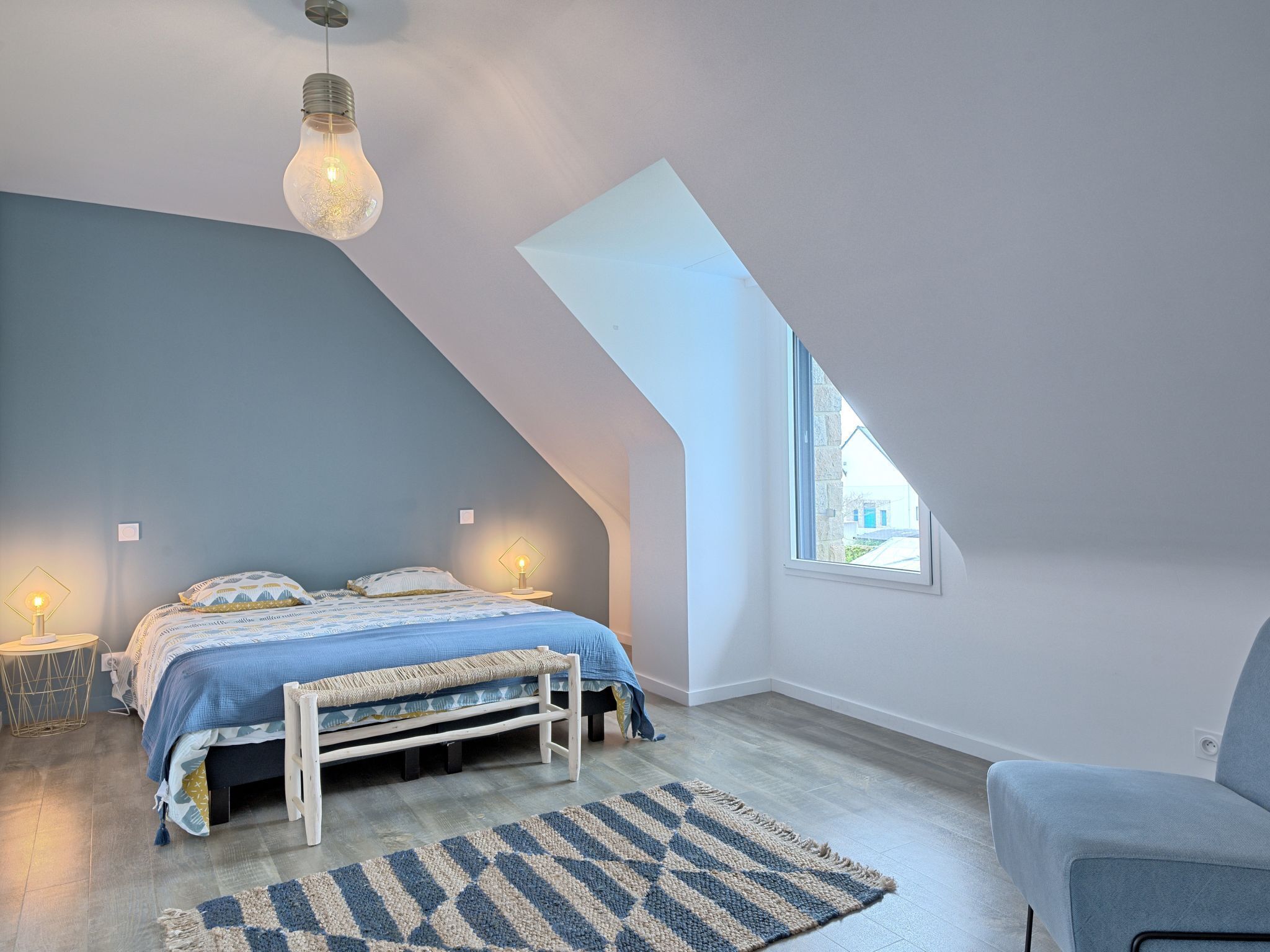 A bedroom with a sloped ceiling, a bed with a blue and white patterned bedspread, a bench, a rug, and a window providing a view outside.