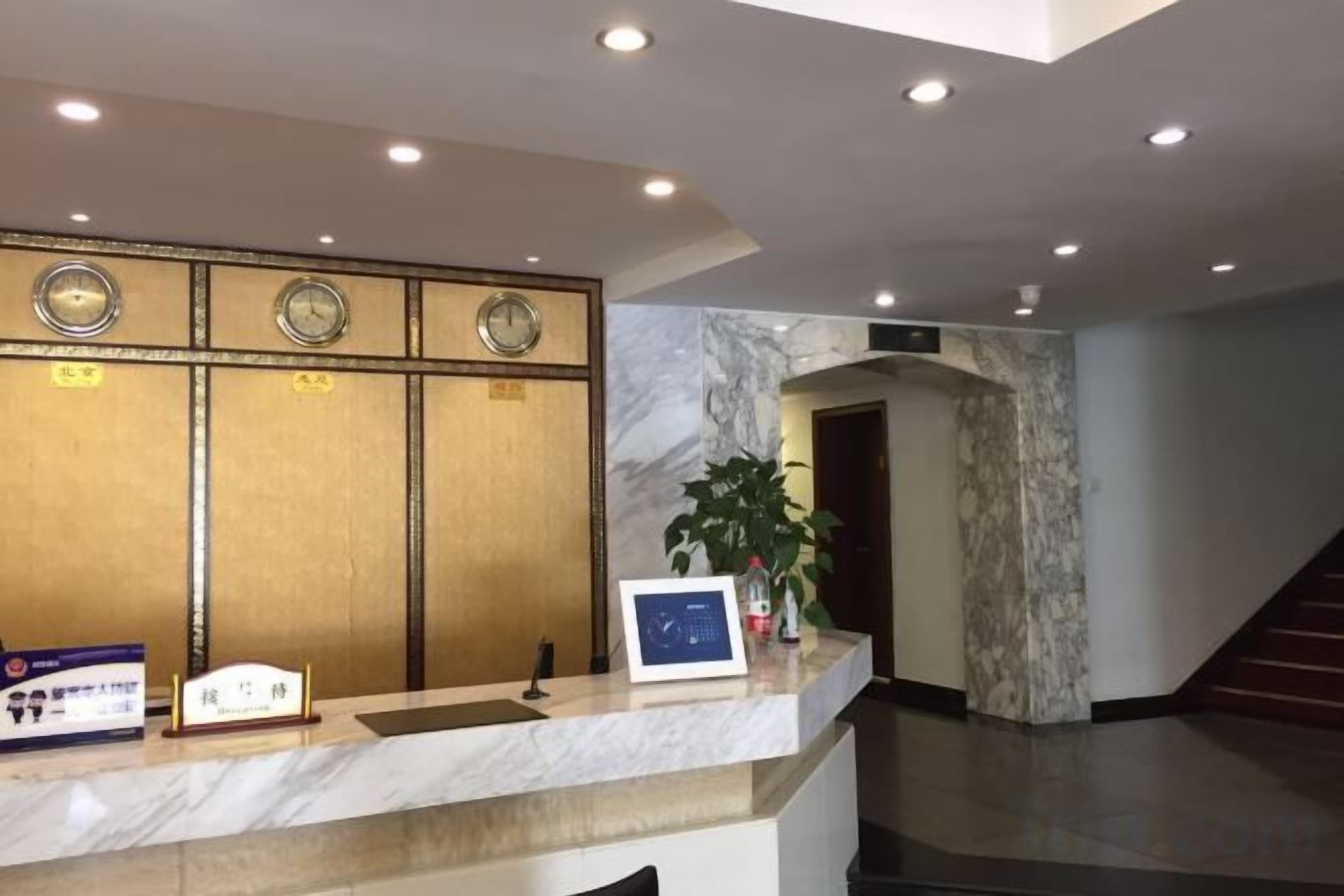 A hotel reception area with a marble counter, clocks, a plant, and a staircase.