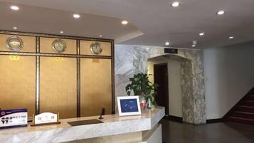 A hotel reception area with a marble counter, clocks, a plant, and a staircase.