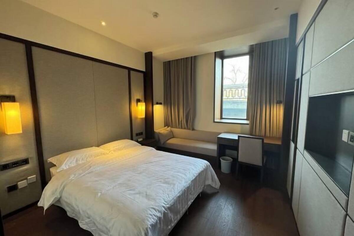 A hotel room with a bed, a desk, a chair, a window with curtains, and a TV mounted on the wall.